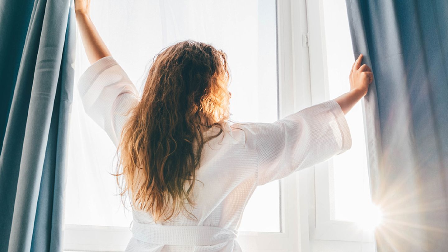 Woman wearing a white robe and opening window curtains to let the sun in