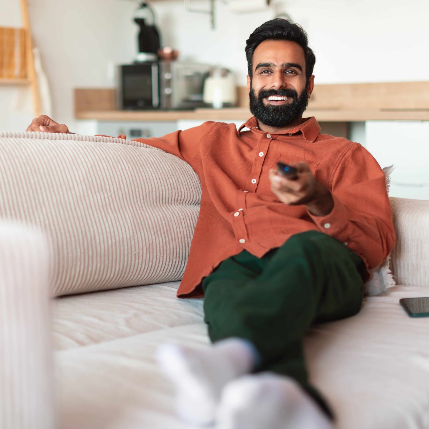 Person relaxing on a couch, smiling while using a TV remote in a modern living room.