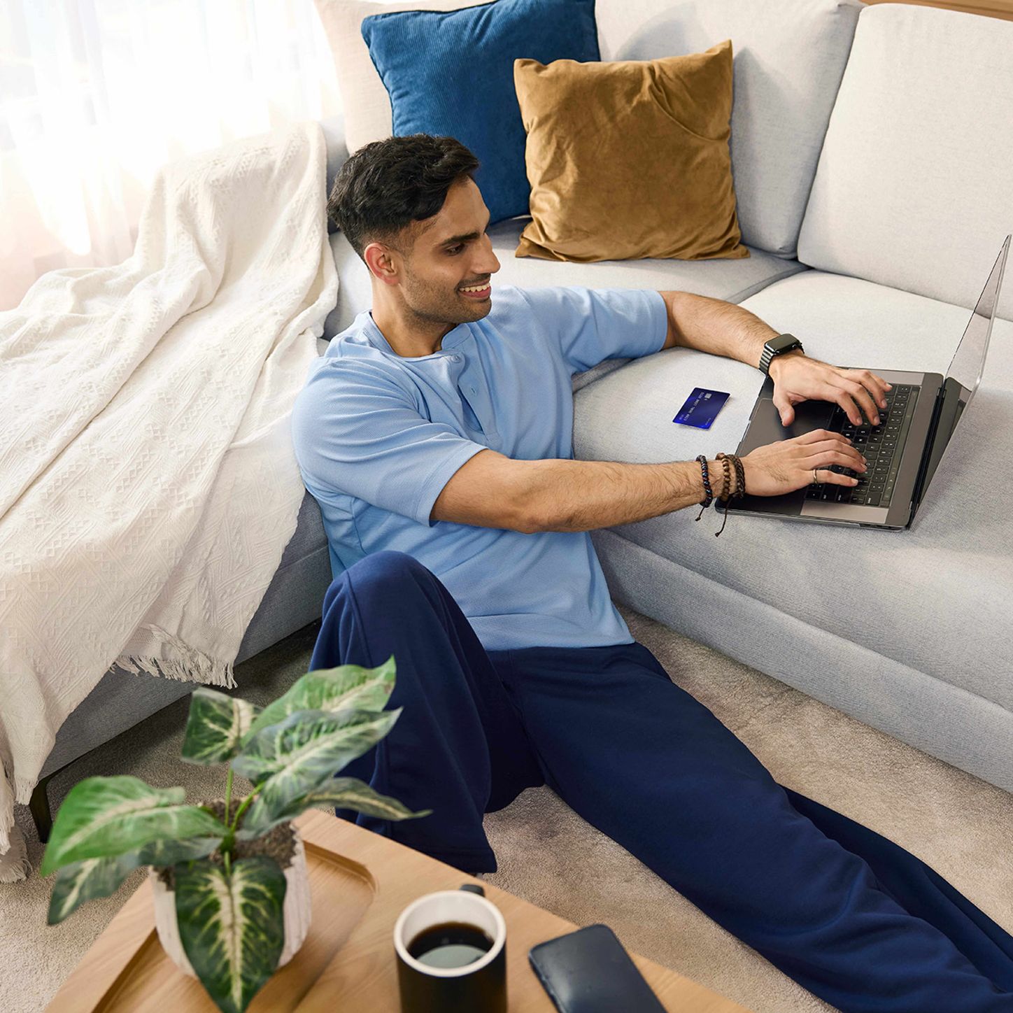 Person sitting on the floor using a laptop while holding a smartphone beside a sofa in a bright living room.