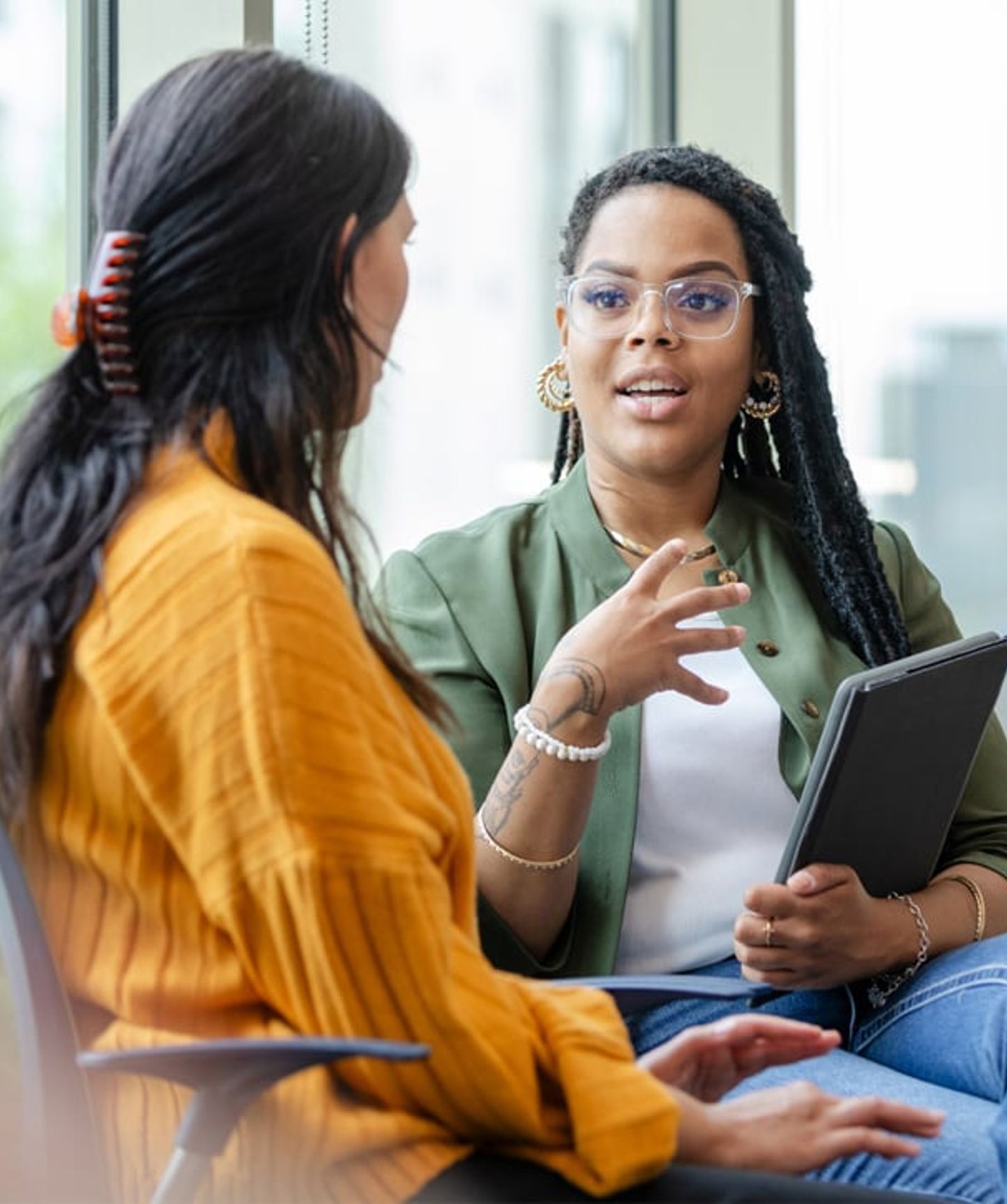 Deux femmes assises discutent dans un bureau moderne avec de grandes fenêtres. L'une d'elles tient une tablette et parle tout en faisant des gestes.