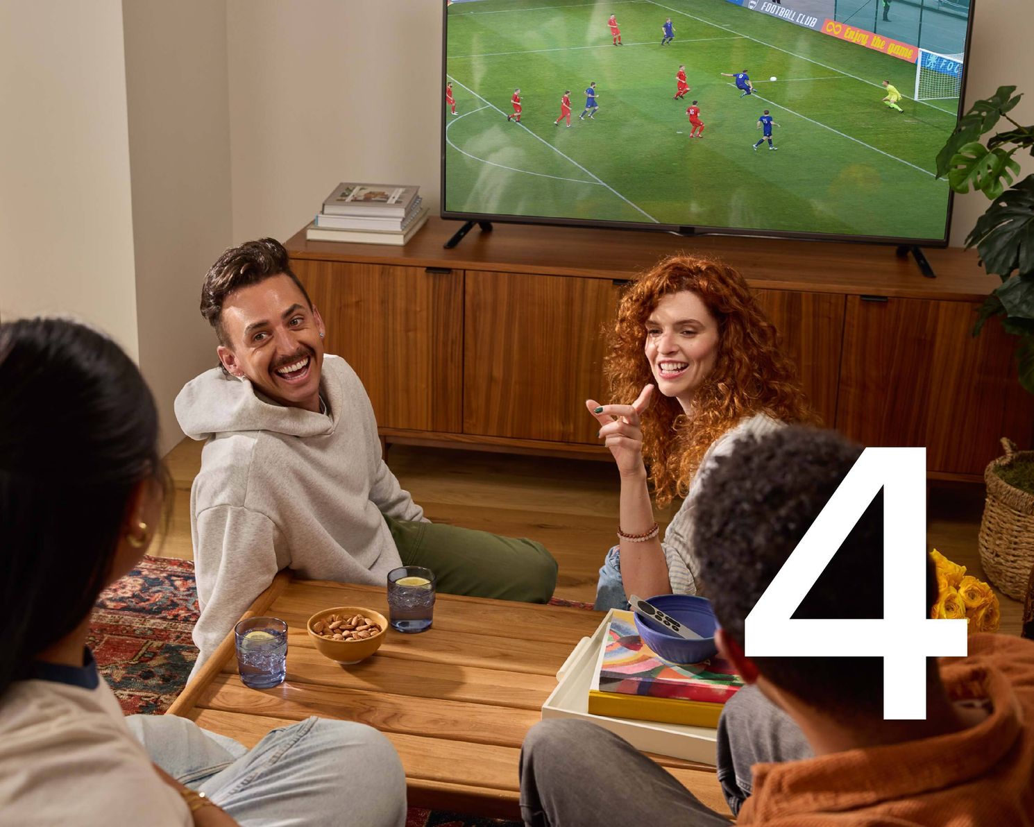 A group of friends sitting on the floor watching a soccer game and laughing with the number 4 overlaid in white text.