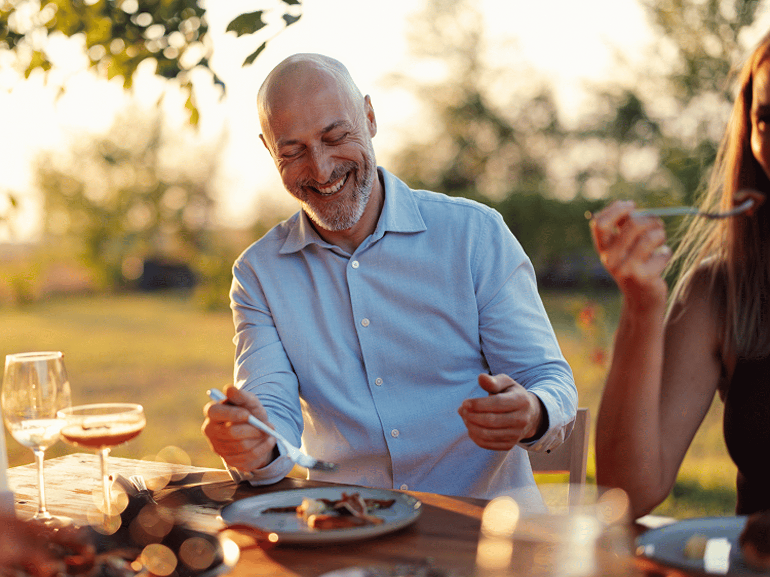 A smiling, middle-aged bald man in a light blue button-down shirt eats dinner outdoors at a wooden table during a warm, golden hour sunset light.