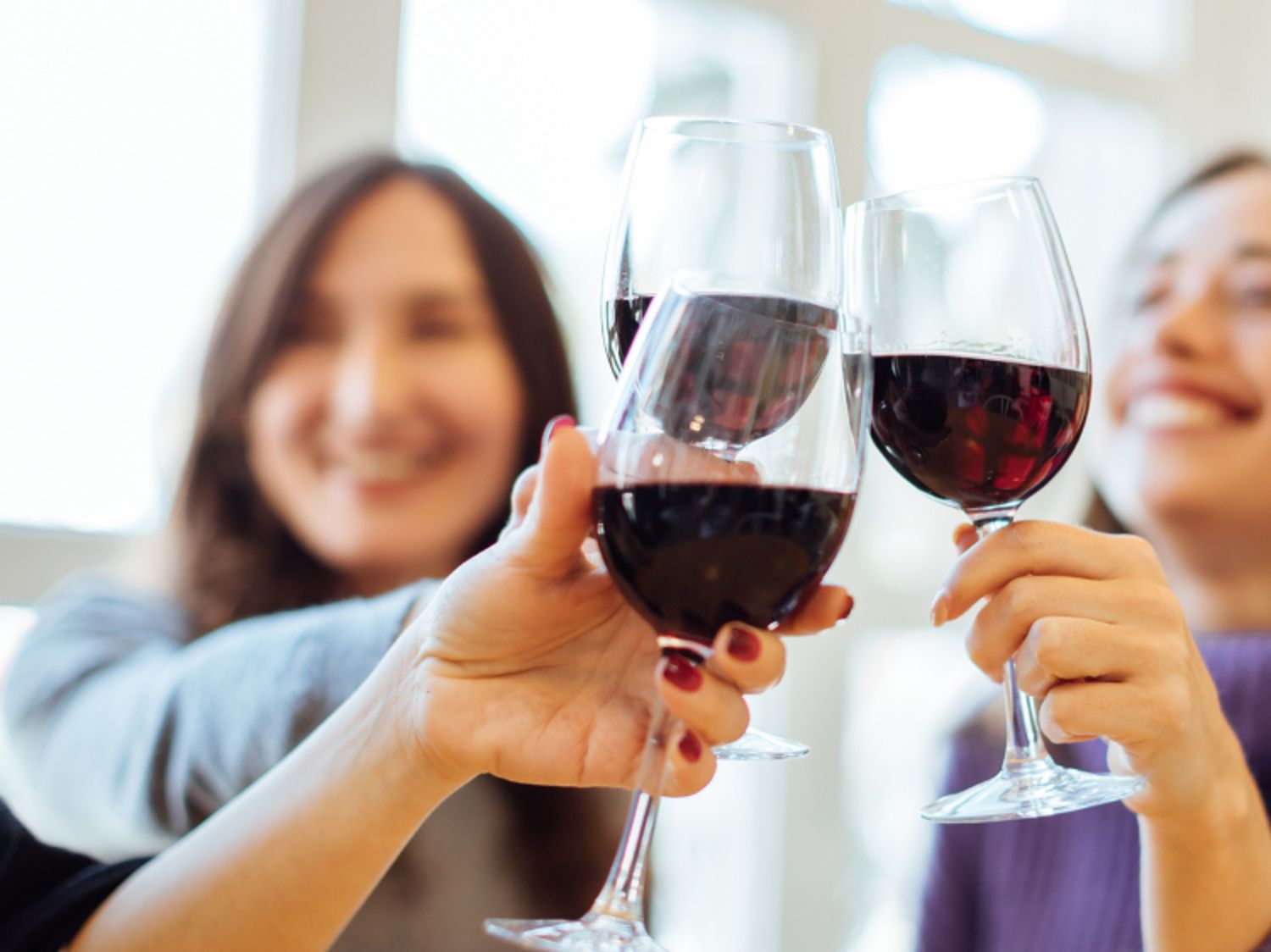 Two women holding clear glass stems filled with deep red wine, clinking their glasses together in a toast.