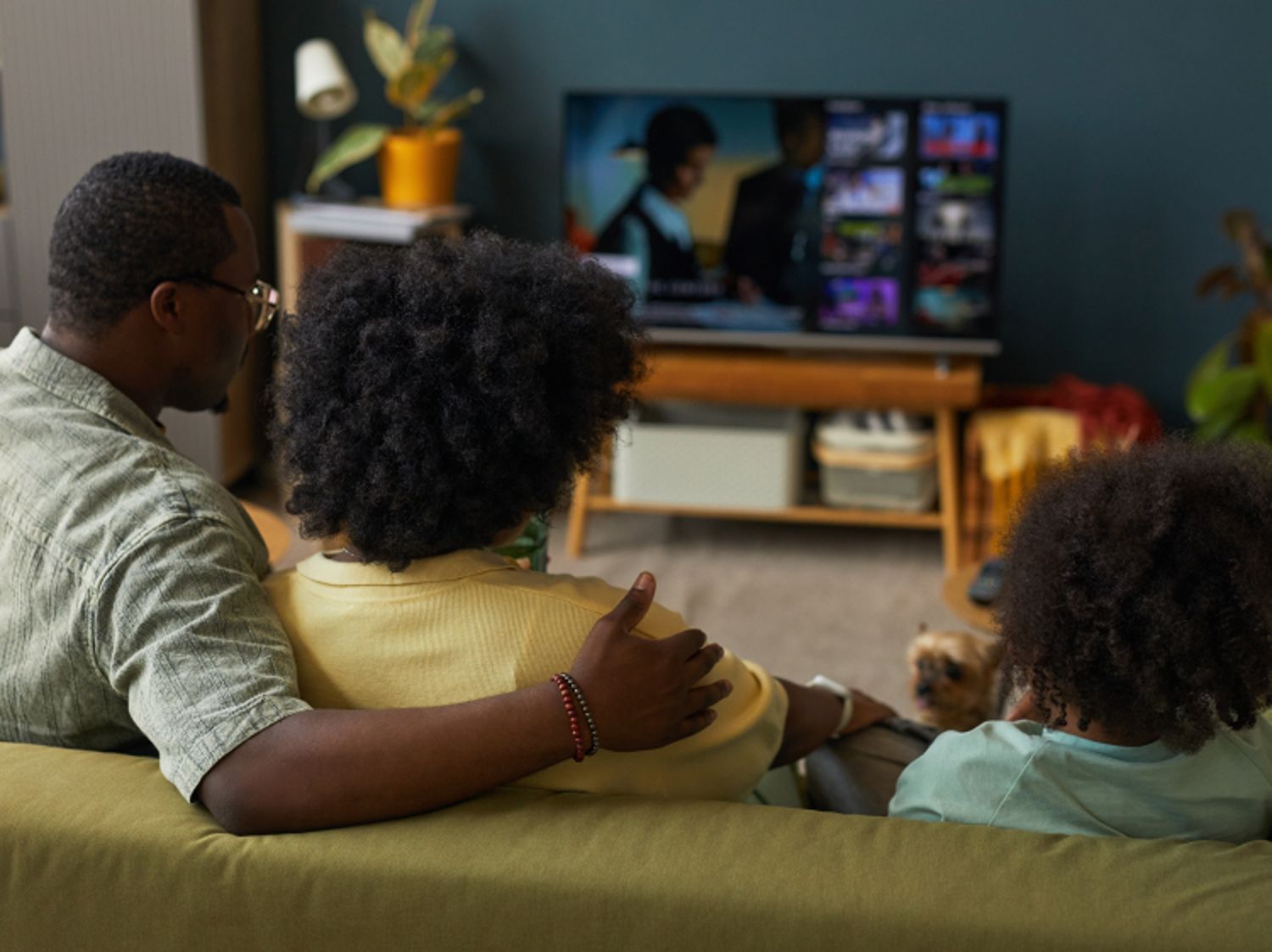 A family sits on the couch watching TV while a small dog sits in the background.