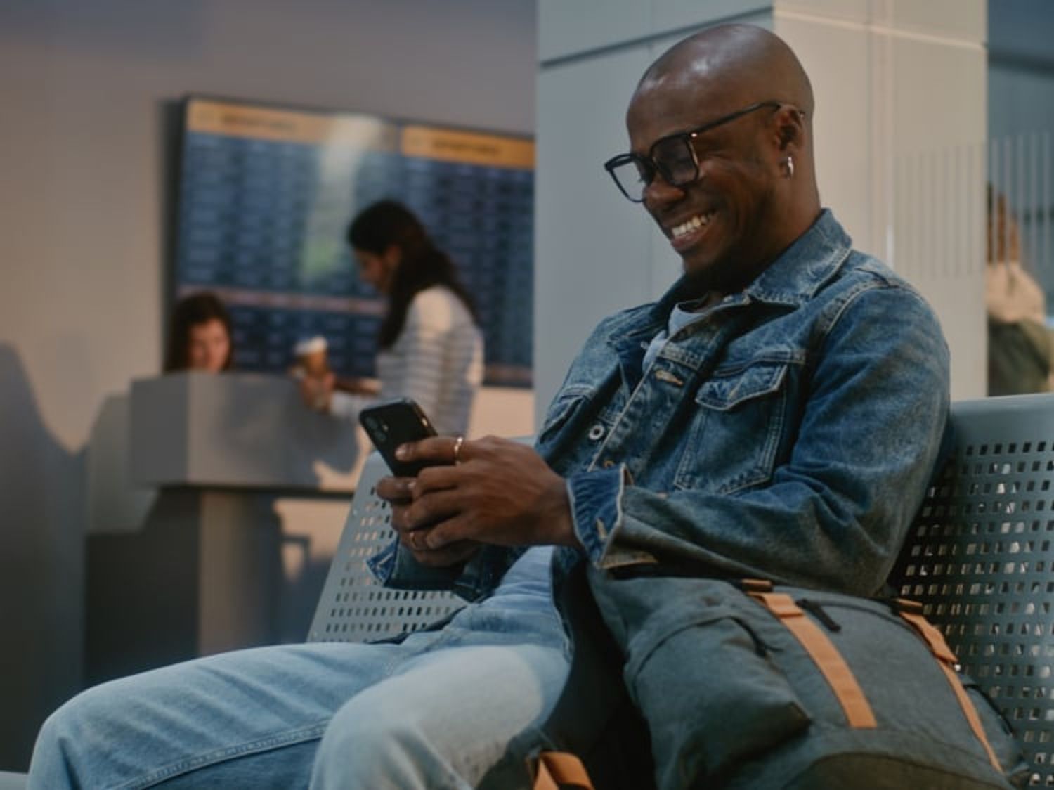 Smiling man wearing glasses and a denim jacket sits on a bench, looking at his phone with a bag beside him in a modern indoor waiting area.
