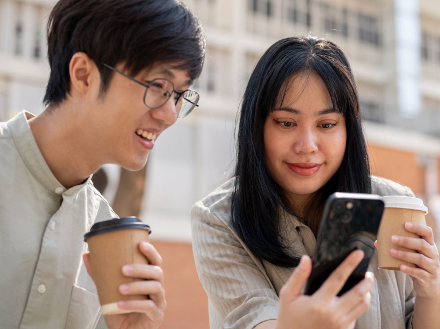 Two people holding coffee cups look at a smartphone together outdoors, with buildings in the background.