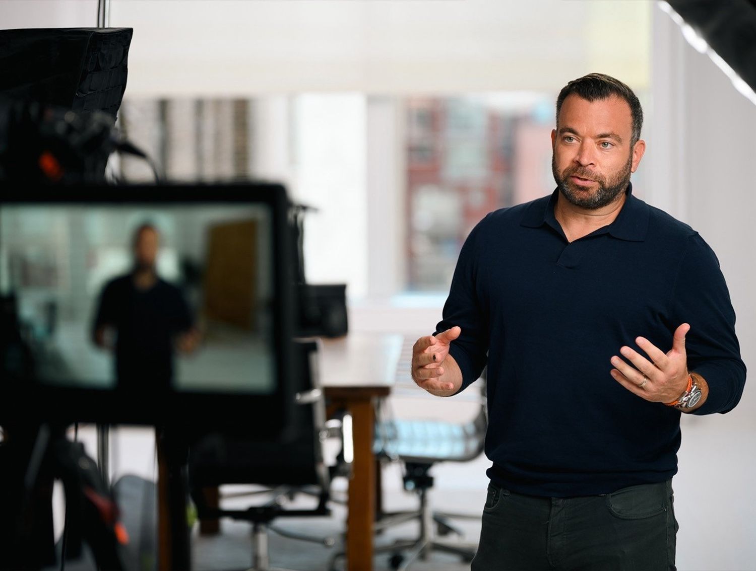 A man in a dark shirt speaks on camera in a bright studio while his image appears on a nearby monitor during filming.