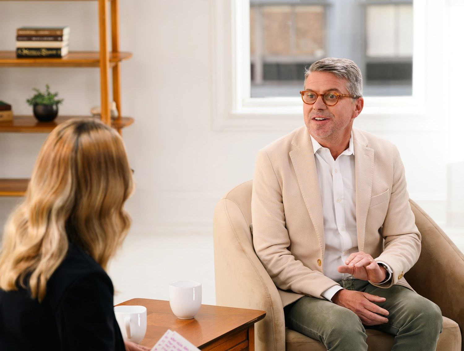 A man in a light blazer speaks with a woman during a relaxed conversation in a bright, modern living-room setting.