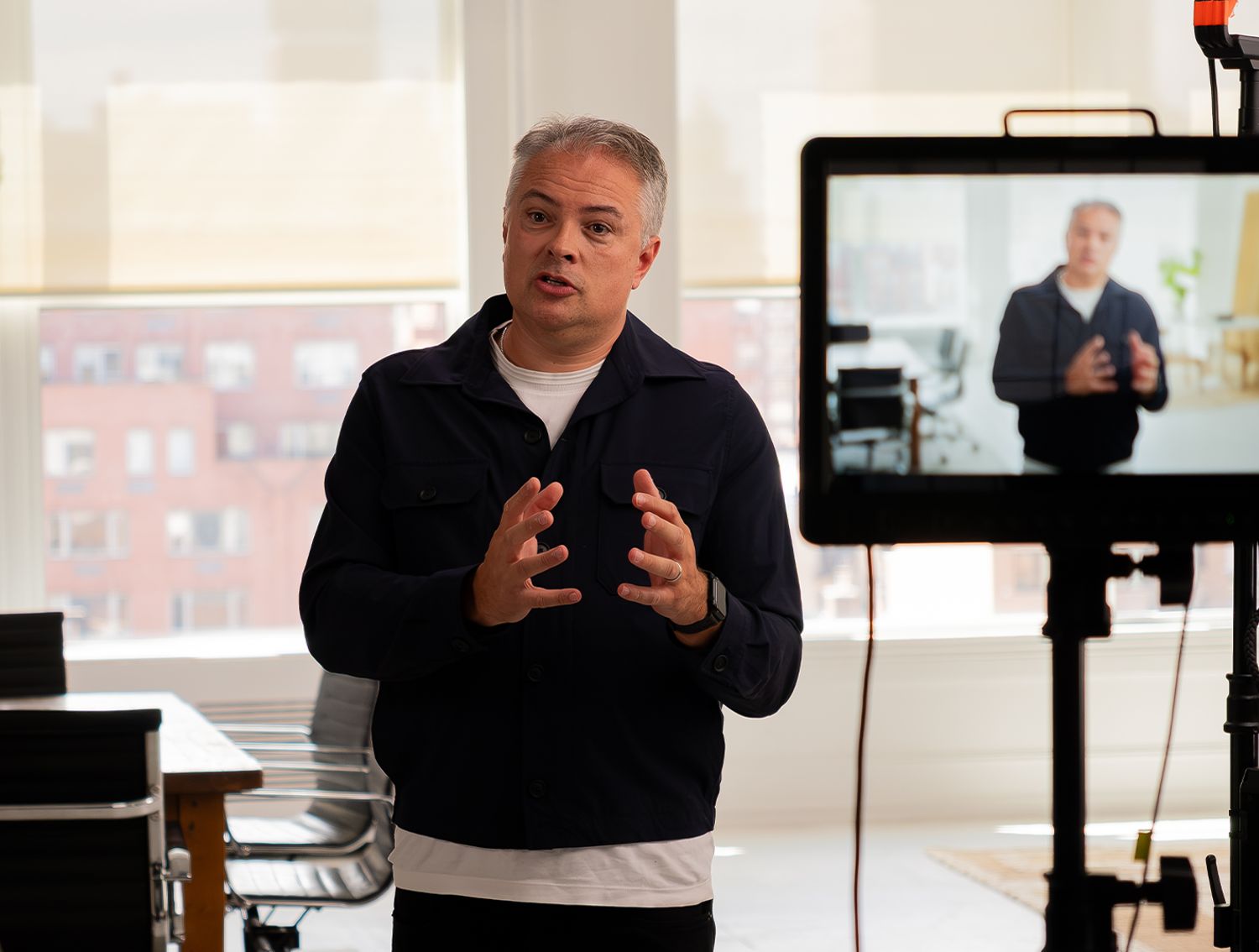 A man speaks to the camera in a bright studio while his live image is displayed on a nearby monitor during a video recording session.