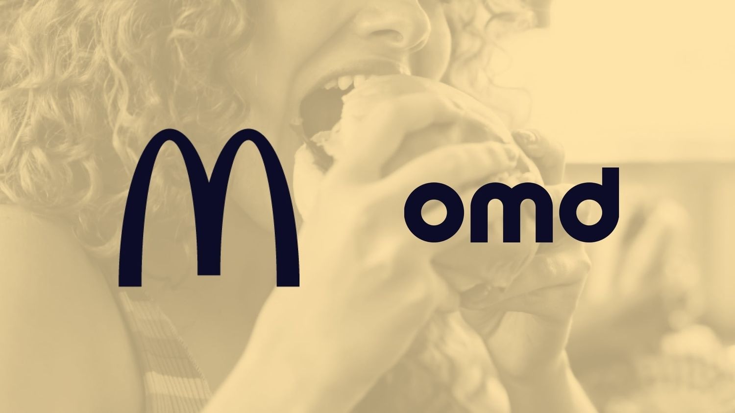 Close-up of a person with curly red hair taking a bite of a sandwich, smiling in a bright, casual setting.