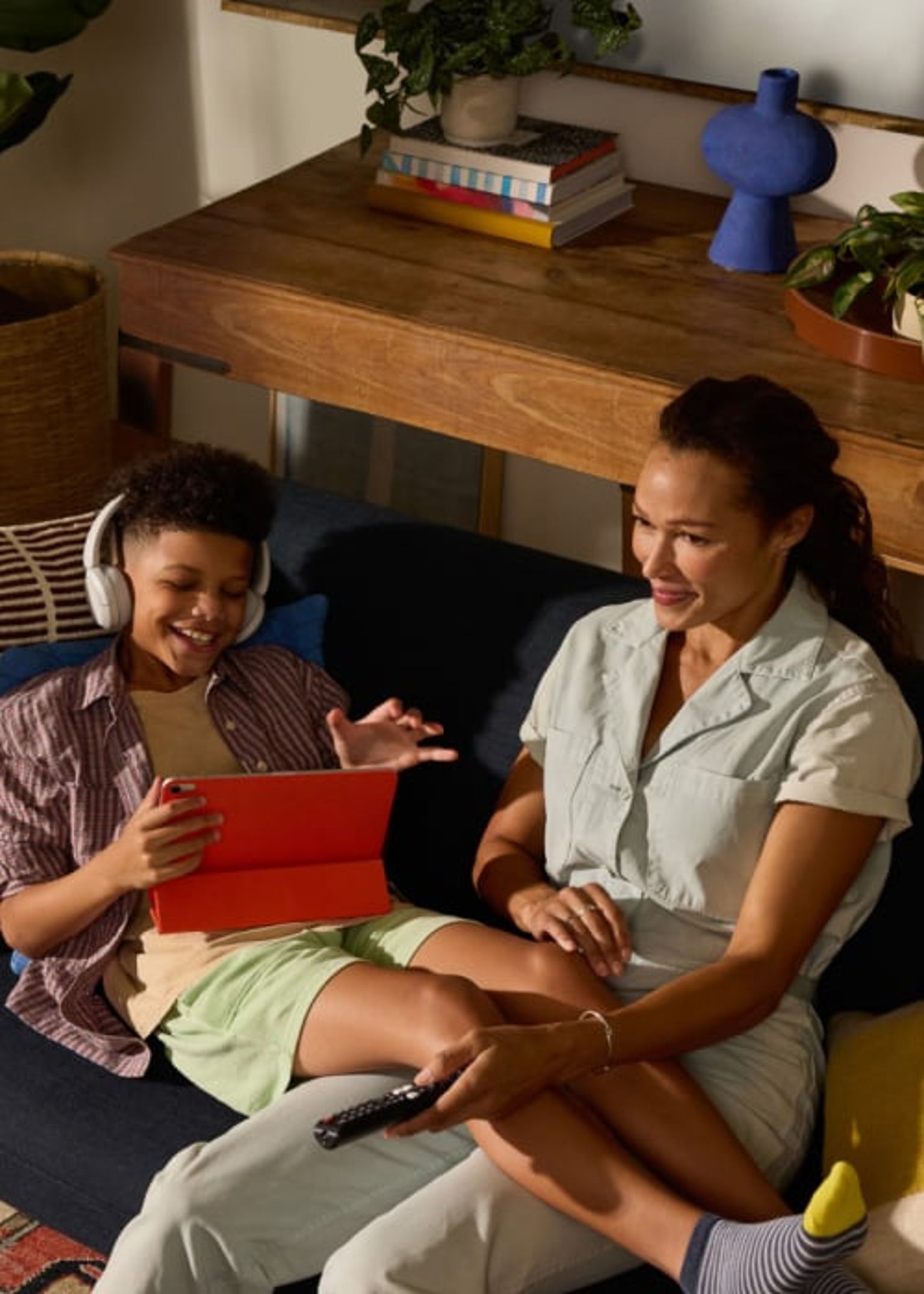 Una mujer y un niño están sentados cómodamente en el sofá de una sala de estar iluminada con calidez, sonriendo y disfrutando de contenido digital en una tableta, el niño lleva auriculares.