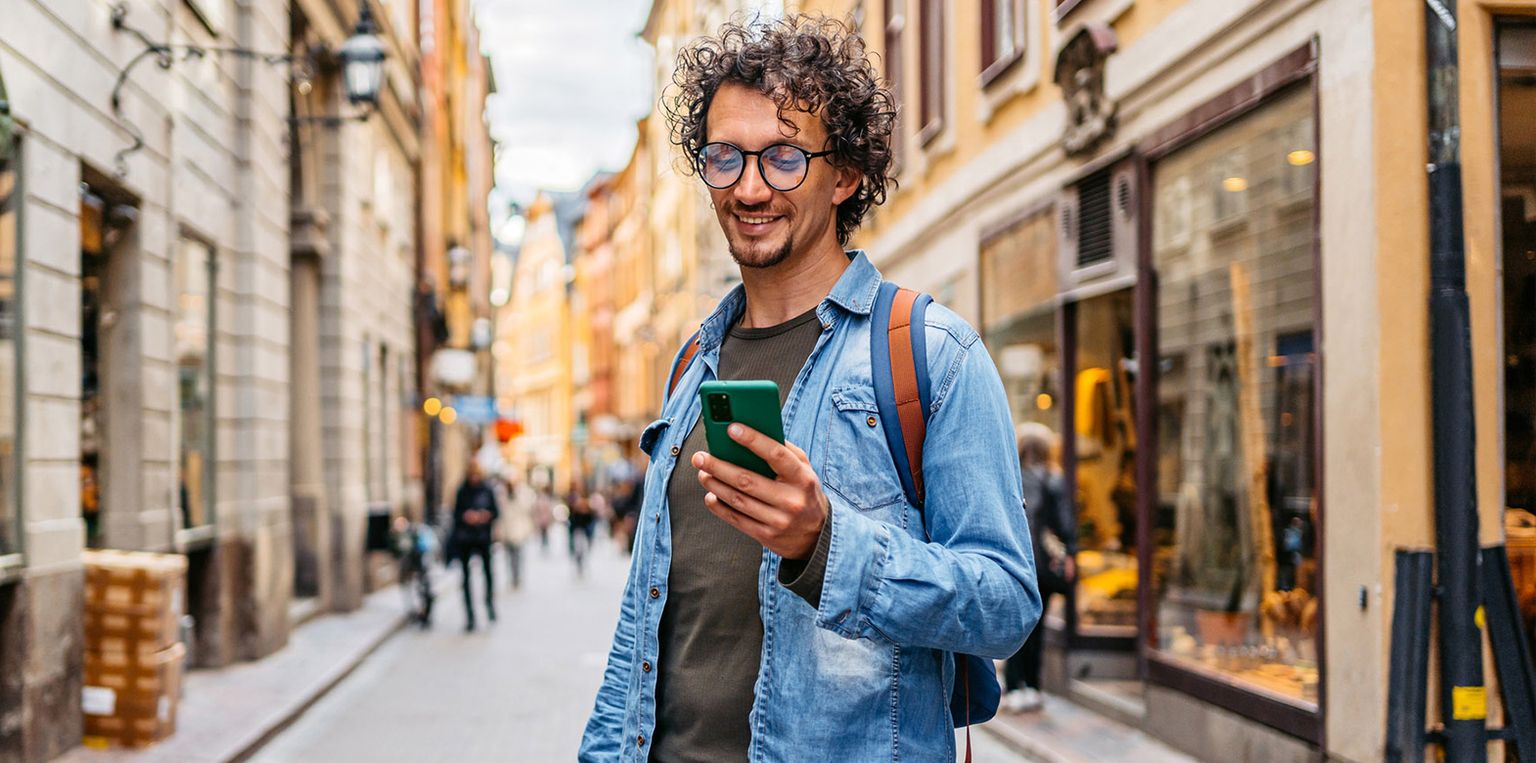 A man with curly hair and glasses smiles while using his smartphone on a vibrant, narrow European street.
