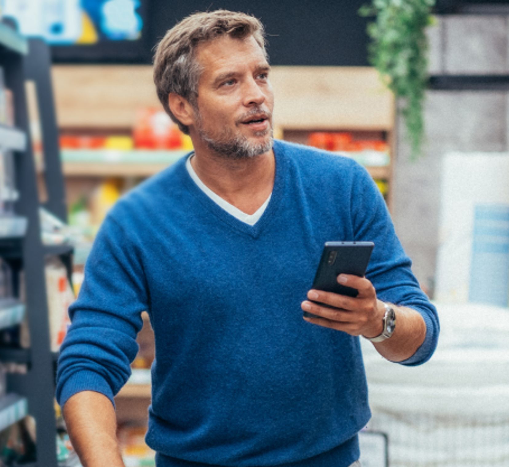 Man in a grocery store holding a smartphone, browsing products while shopping in an aisle with shelves on both sides.