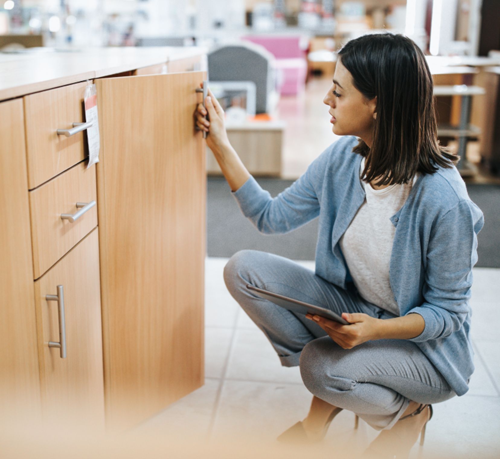 Woman kneeling in a kitchen aisle, using a tablet while checking cabinet products, comparing options before purchase.