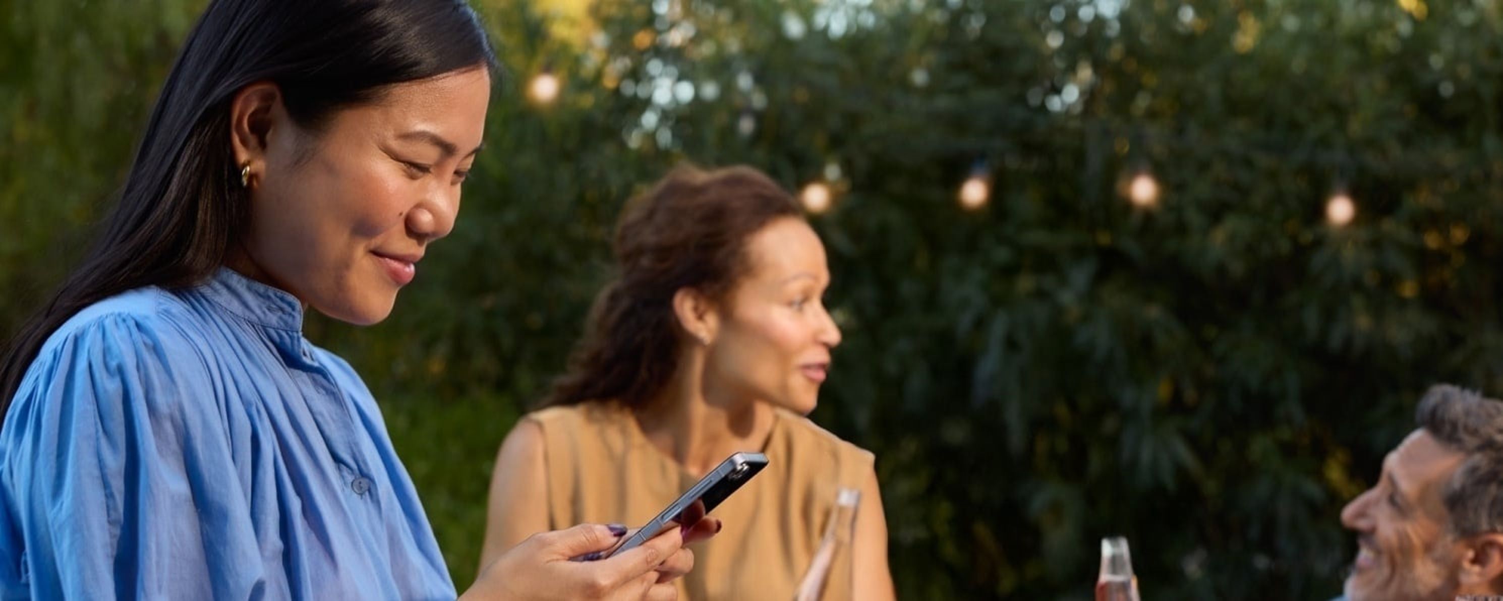 A woman in a blue blouse smiles while looking at her phone during an outdoor gathering with friends.