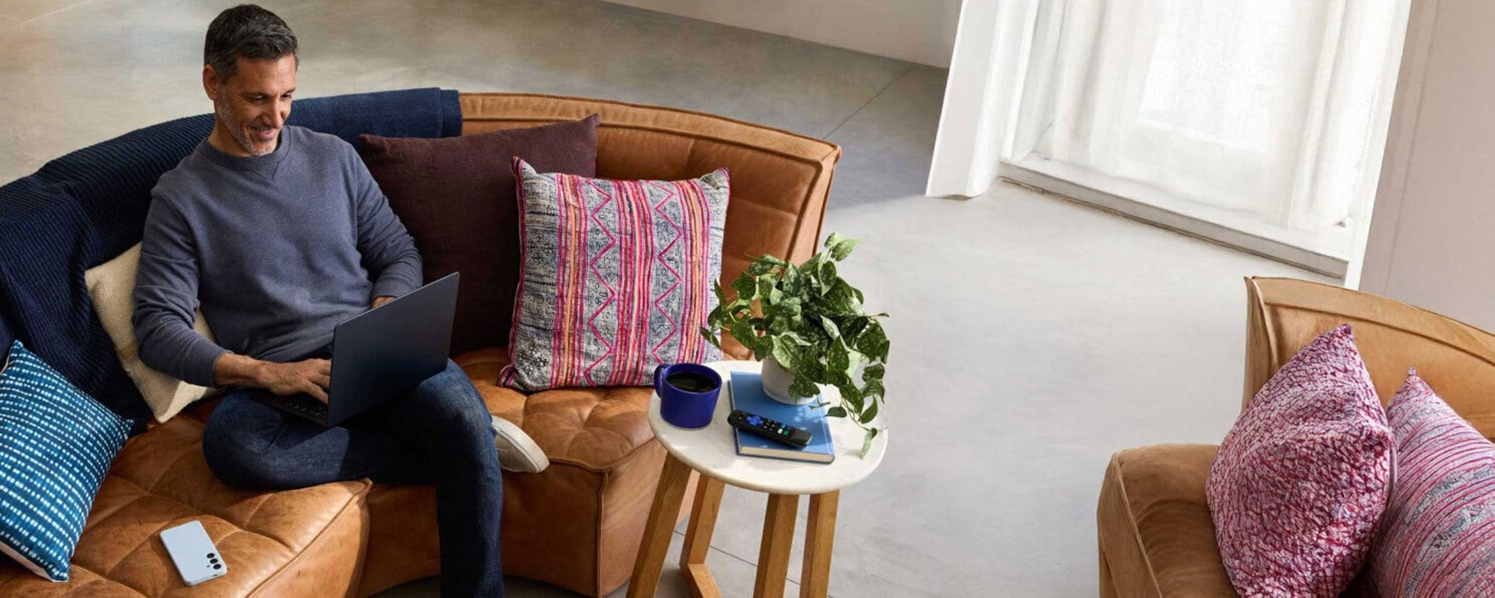 A man sits comfortably on a couch with colorful pillows, smiling while working on a laptop.