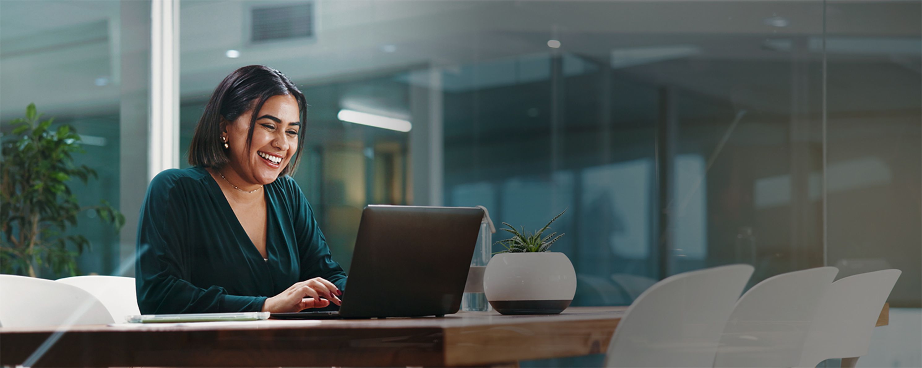A woman in a green blouse smiles while looking at her laptop in an office setting.