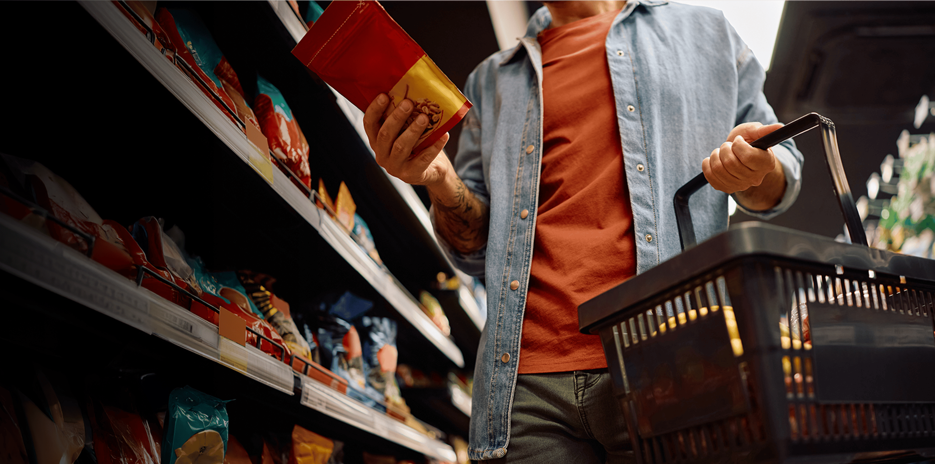 Person shopping in a grocery aisle, holding a snack bag and basket, comparing products before making a purchase.