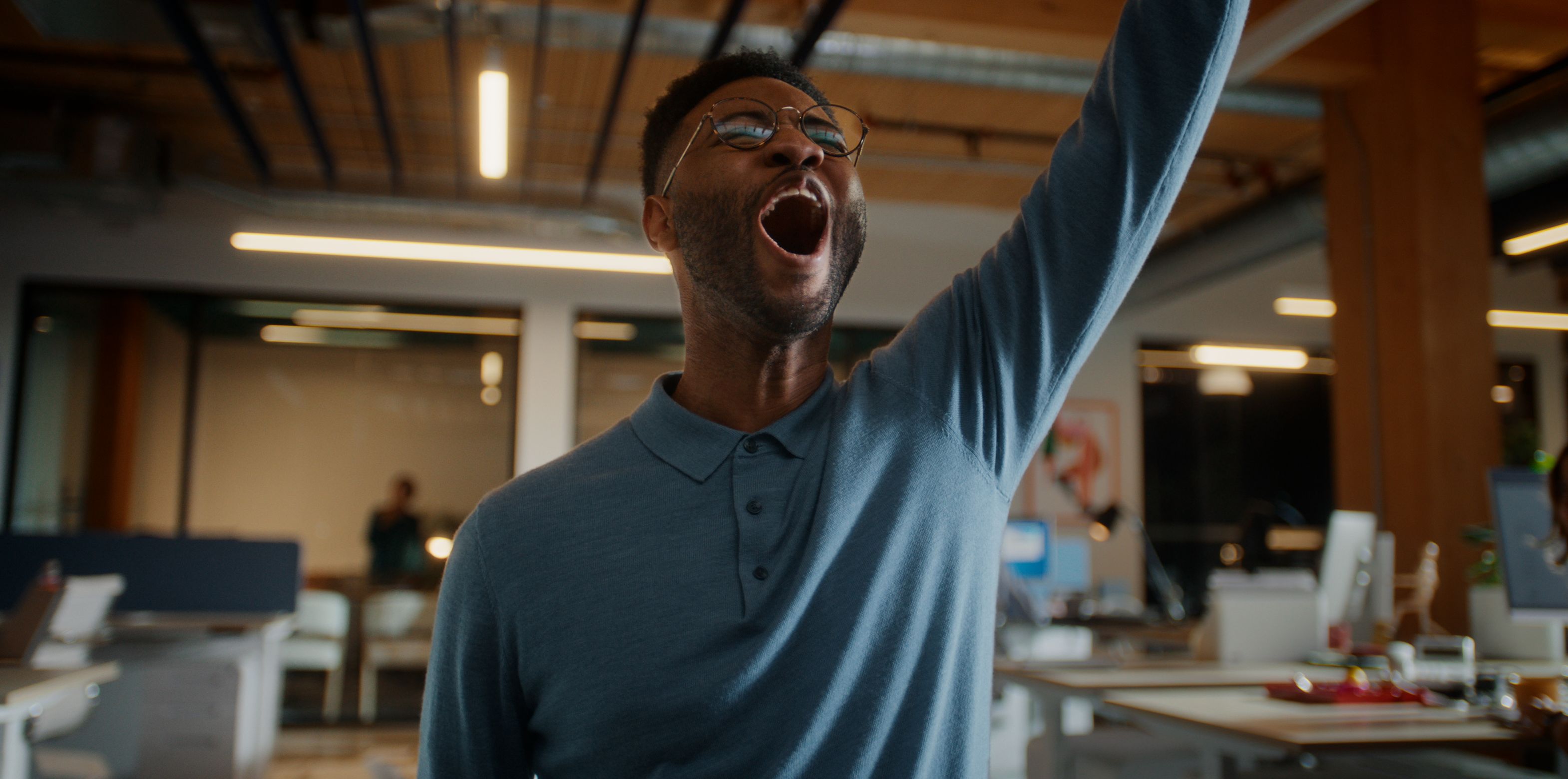 Person standing in a modern office with one arm raised and mouth open, with desks and computers visible in the background.