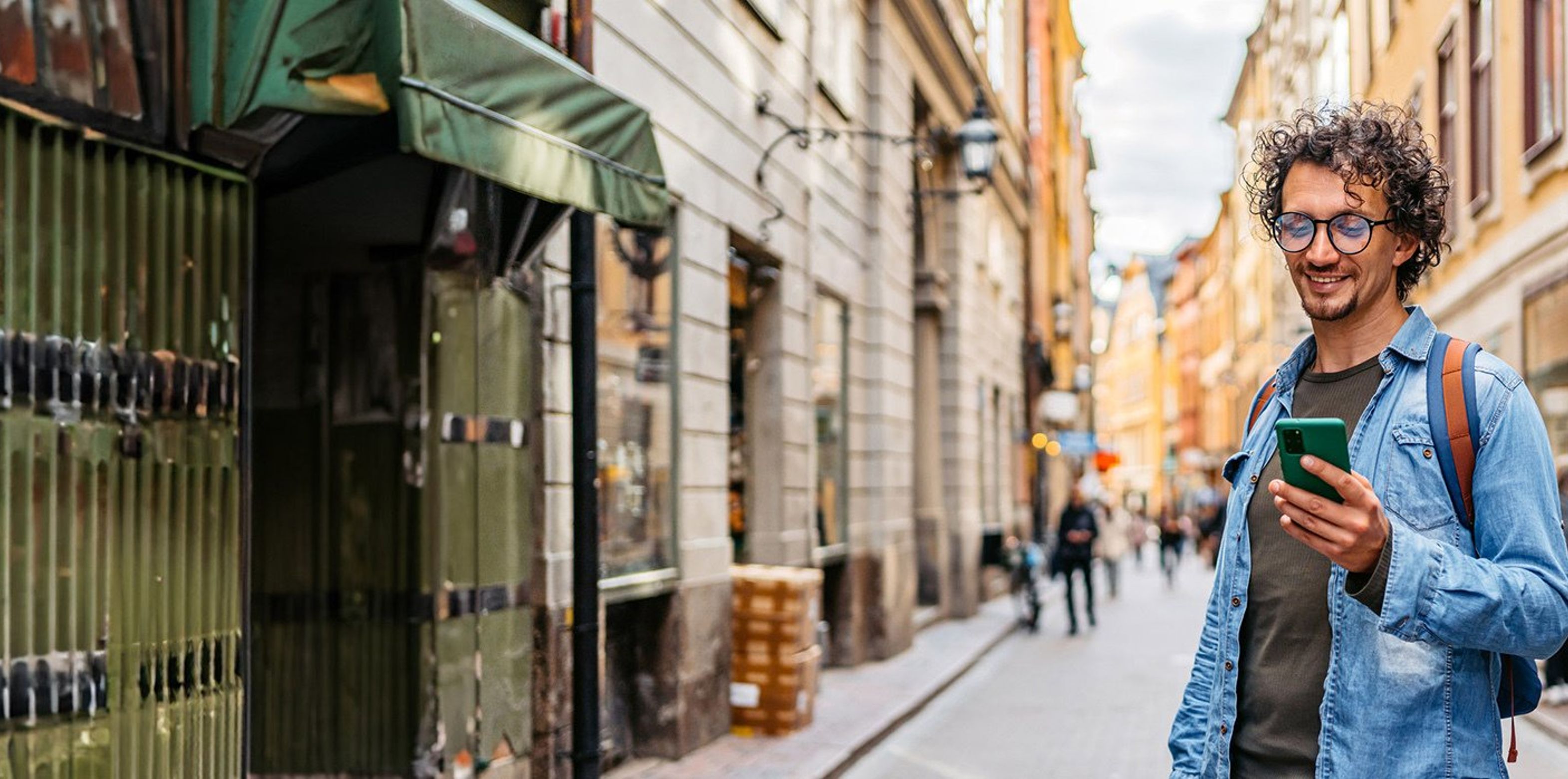 A man with curly hair and glasses smiles while using his smartphone on a vibrant, narrow European street.