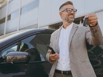 Smiling man in a beige blazer holding car keys and a smartphone while standing next to a black car outside a modern building.