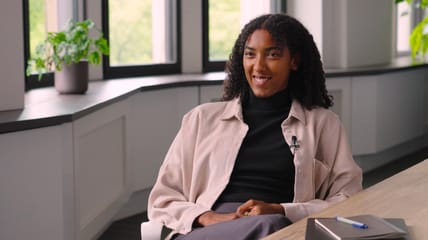 A woman sits at a desk in a modern office setting, speaking on camera