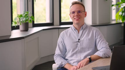 Eine lächelnde Person mit Brille und hellblauem Hemd sitzt an einem Tisch in einem hellen, modernen Büro mit Pflanzen und großen Fenstern im Hintergrund.