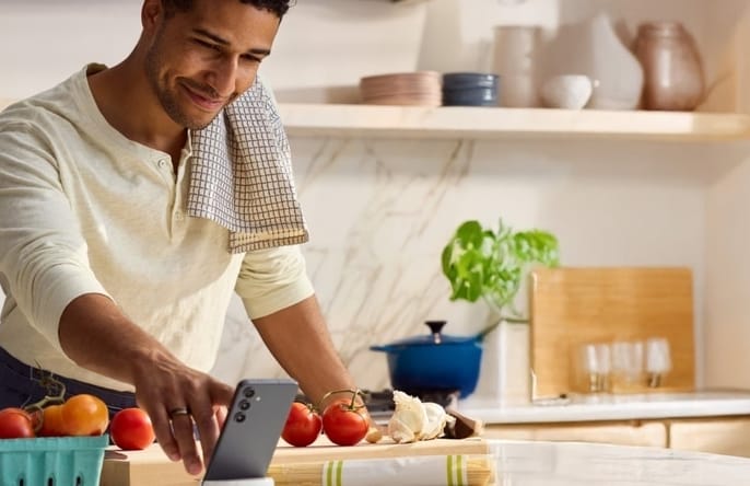 A man in a kitchen with a towel over his shoulder smiles while using his phone near fresh vegetables.