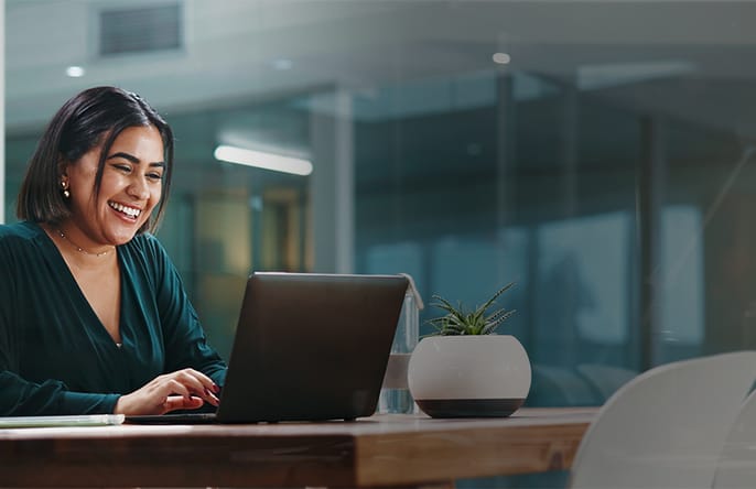 A woman in a green blouse smiles while looking at her laptop in an office setting.