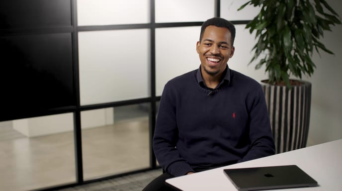 Man smiling in an office setting with a laptop on the desk and a plant in the background.