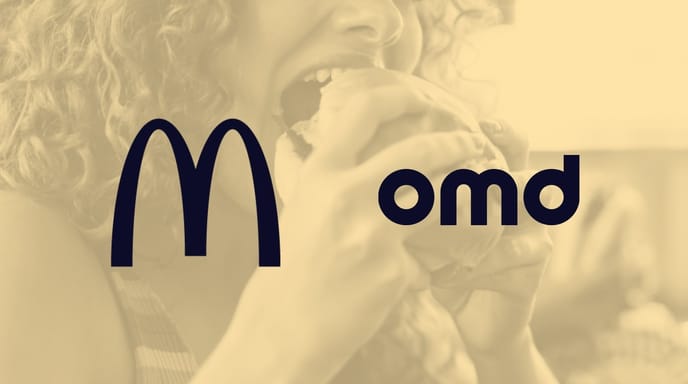 Close-up of a person with curly red hair taking a bite of a sandwich, smiling in a bright, casual setting.
