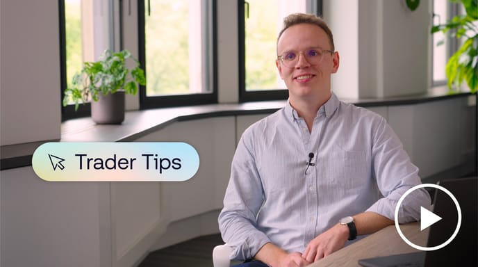 A smiling person wearing glasses and a light blue shirt sits in a bright modern office with plants and large windows, with a “Trader Tips” graphic and a play button icon on screen.