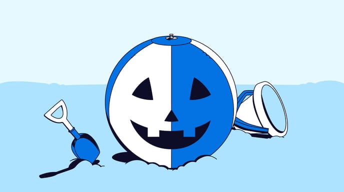 A blue and white beach ball designed as a Halloween pumpkin, partially submerged in sand with a shovel and bucket nearby.