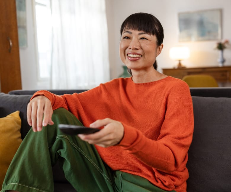 Smiling woman in an orange sweater sitting on a couch holding a TV remote in a cozy living room.