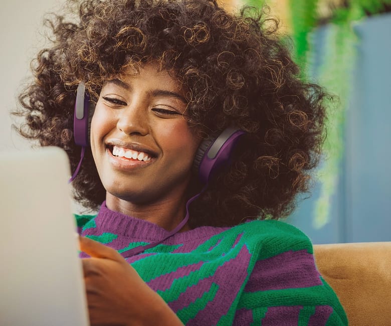 A woman wearing headphones smiles as she watches something on her laptop in a cozy indoor setting.