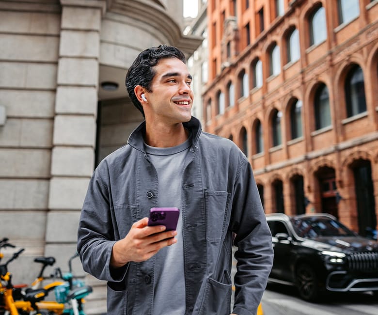 A man smiles while walking through a city street, holding his phone and wearing wireless earbuds against a backdrop of historic buildings.