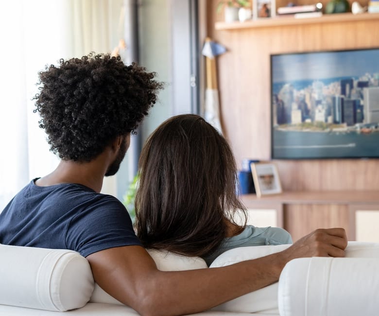 A couple relaxes together on a couch, watching a large screen displaying a cityscape in a cozy living room.