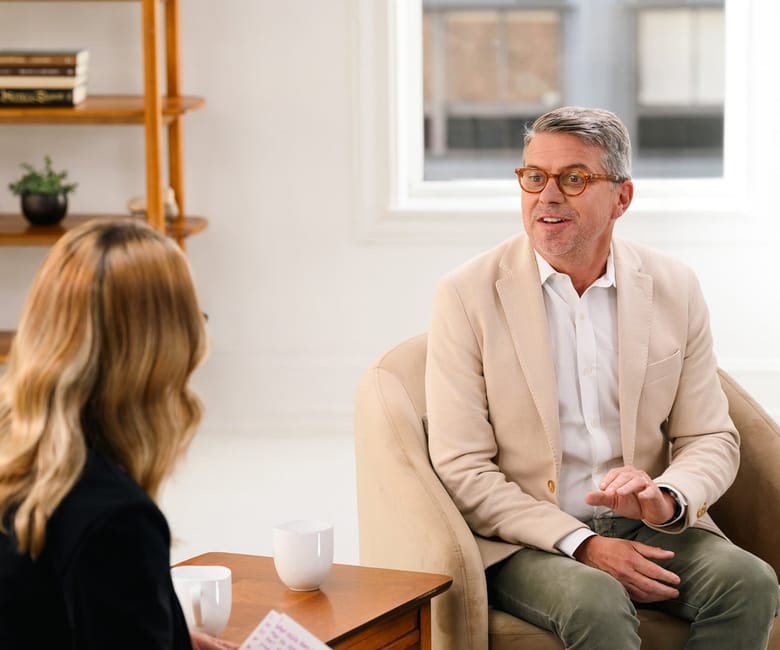 A man in a light blazer speaks with a woman during a relaxed conversation in a bright, modern living-room setting.