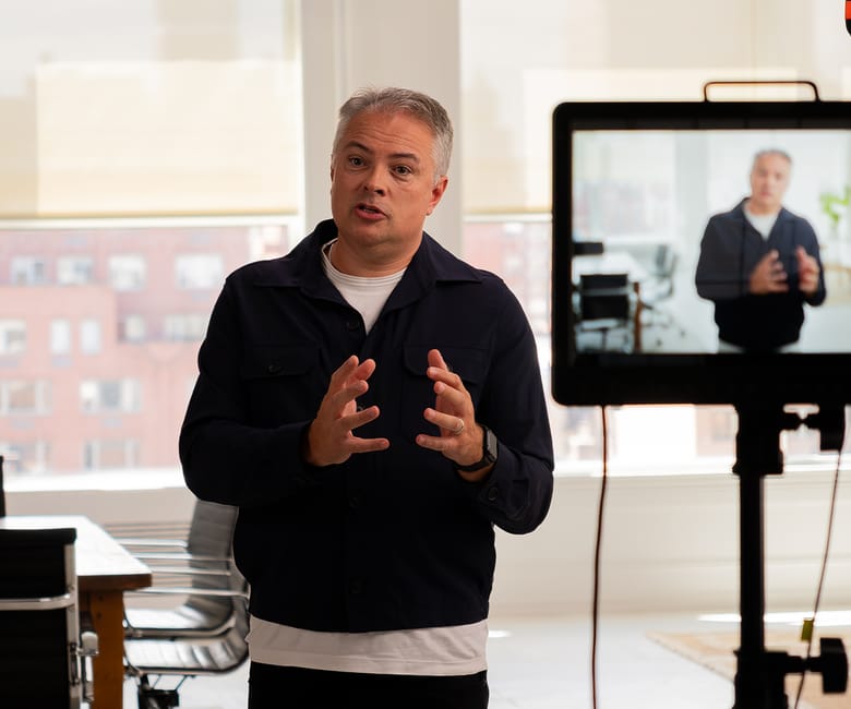 A man speaks to the camera in a bright studio while his live image is displayed on a nearby monitor during a video recording session.