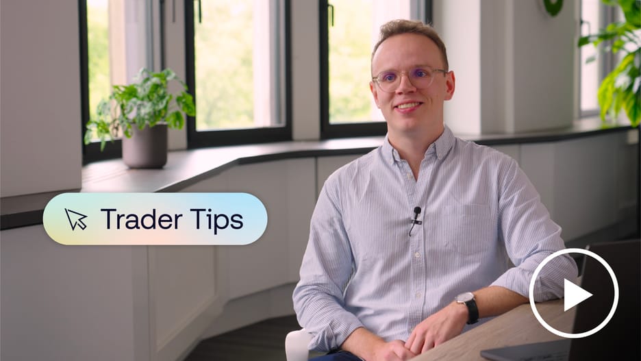 A smiling person wearing glasses and a light blue shirt sits in a bright modern office with plants and large windows, with a “Trader Tips” graphic and a play button icon on screen.