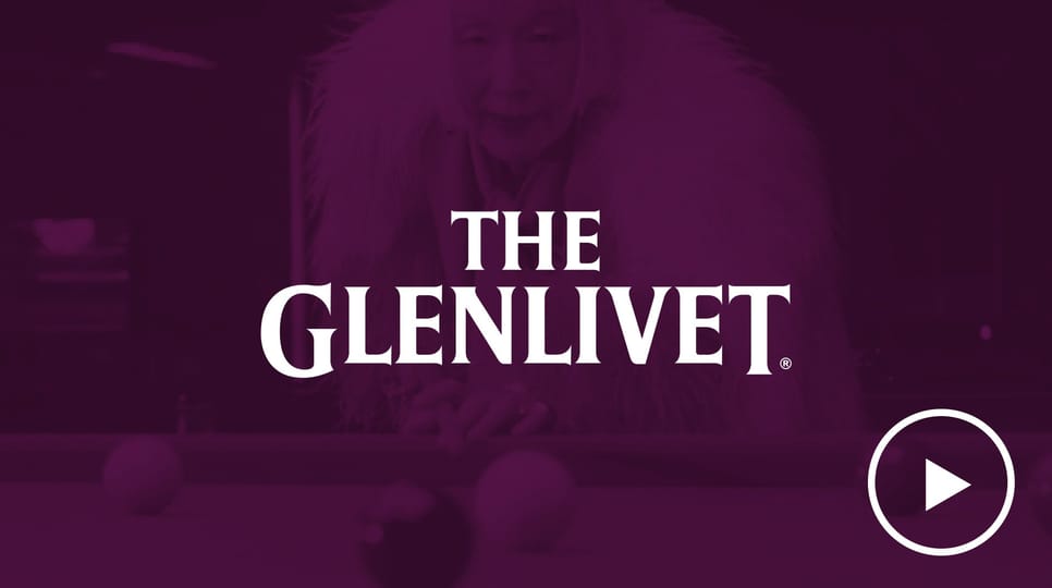 An older woman in a bold fur coat leans over a pool table, overlaid with a purple filter and The Glenlivet logo centered above a play button icon.