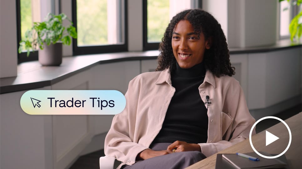 A woman sits at a desk in a modern office setting, speaking on camera