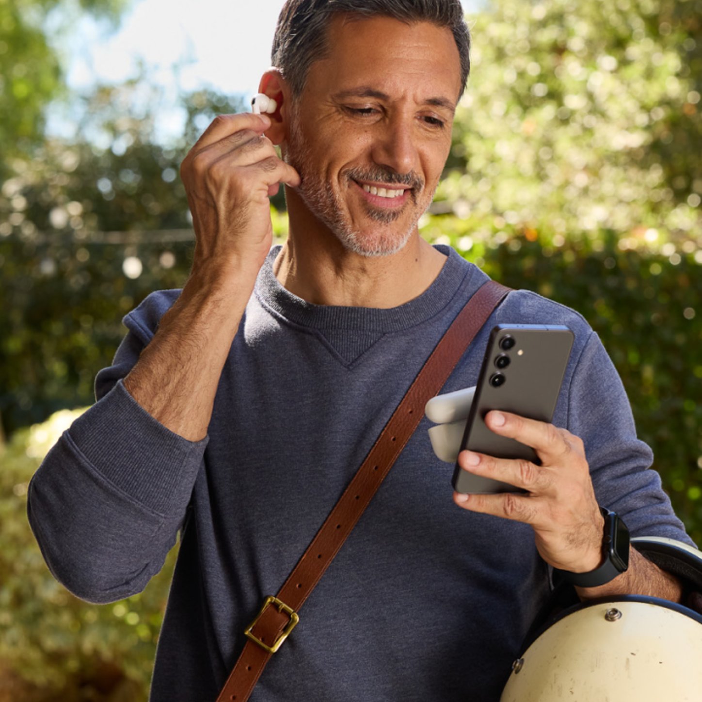 A smiling man outdoors holds a smartphone and inserts a wireless earbud, suggesting he's ready to listen to audio content while on the go.