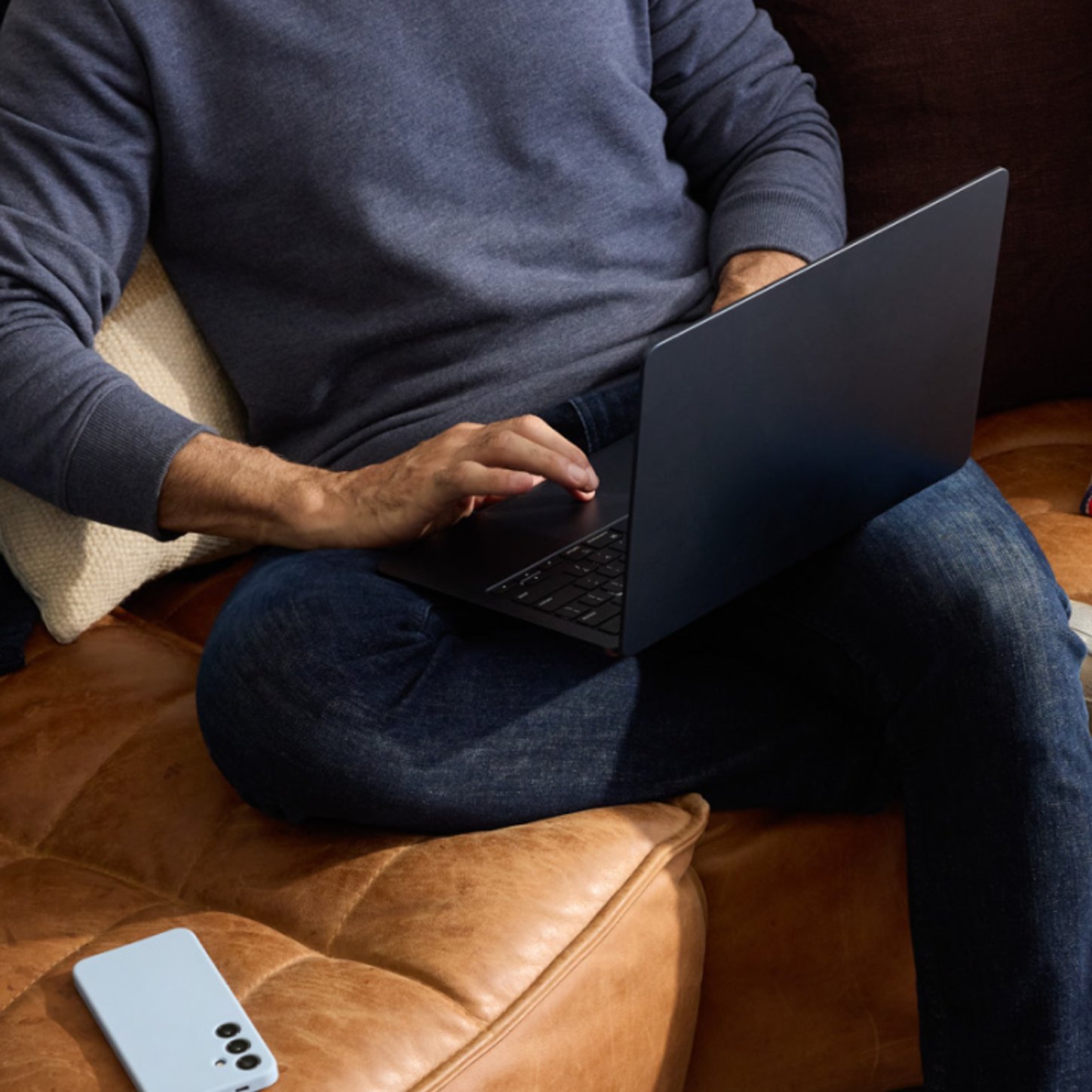 A person sits comfortably on a leather couch using a laptop, with a smartphone placed nearby—conveying a relaxed, tech-savvy work or browsing environment.
