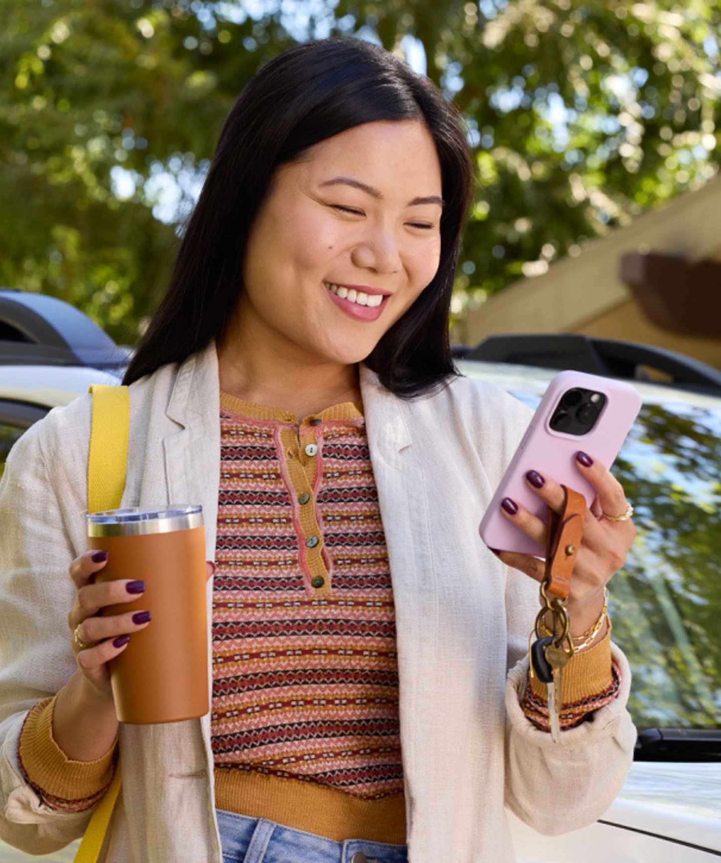 Smiling woman standing outdoors with a coffee tumbler and smartphone, checking her phone near a parked car