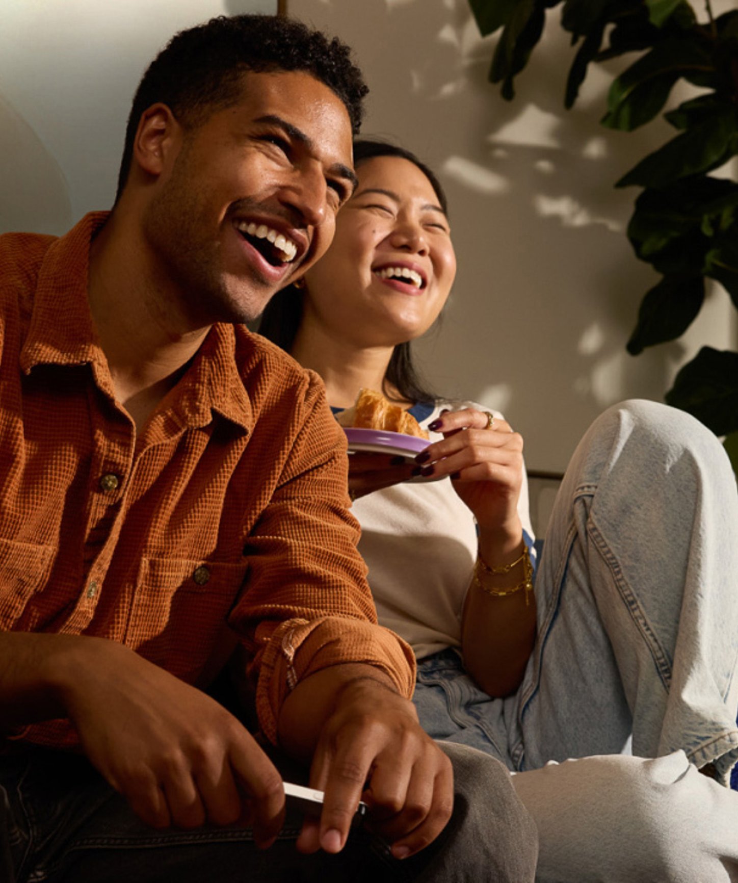 A happy couple laughing while watching TV, sitting on a cozy couch with snacks in hand