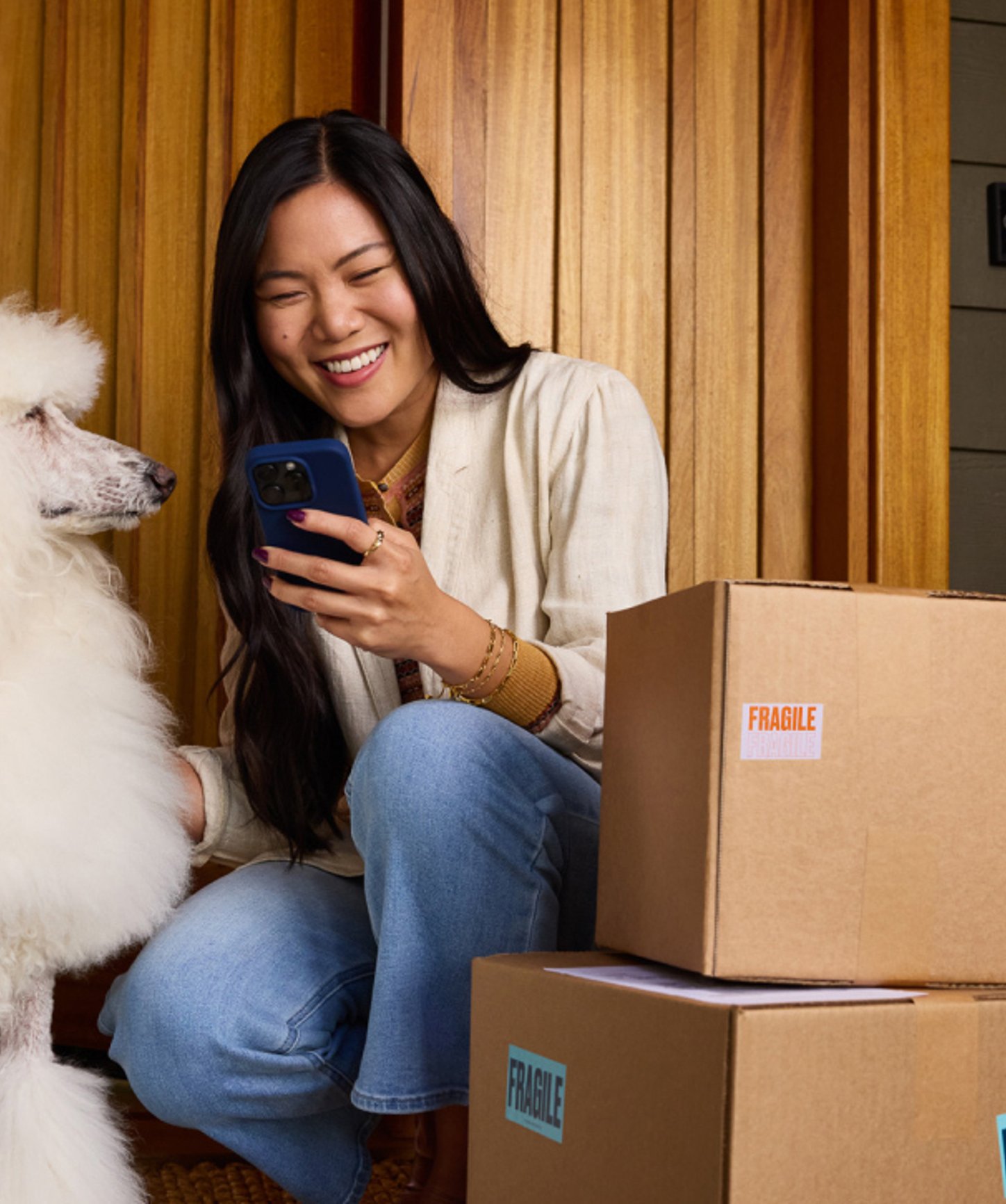 A woman smiling while checking her phone, sitting next to her fluffy white poodle and some fragile-labeled packages