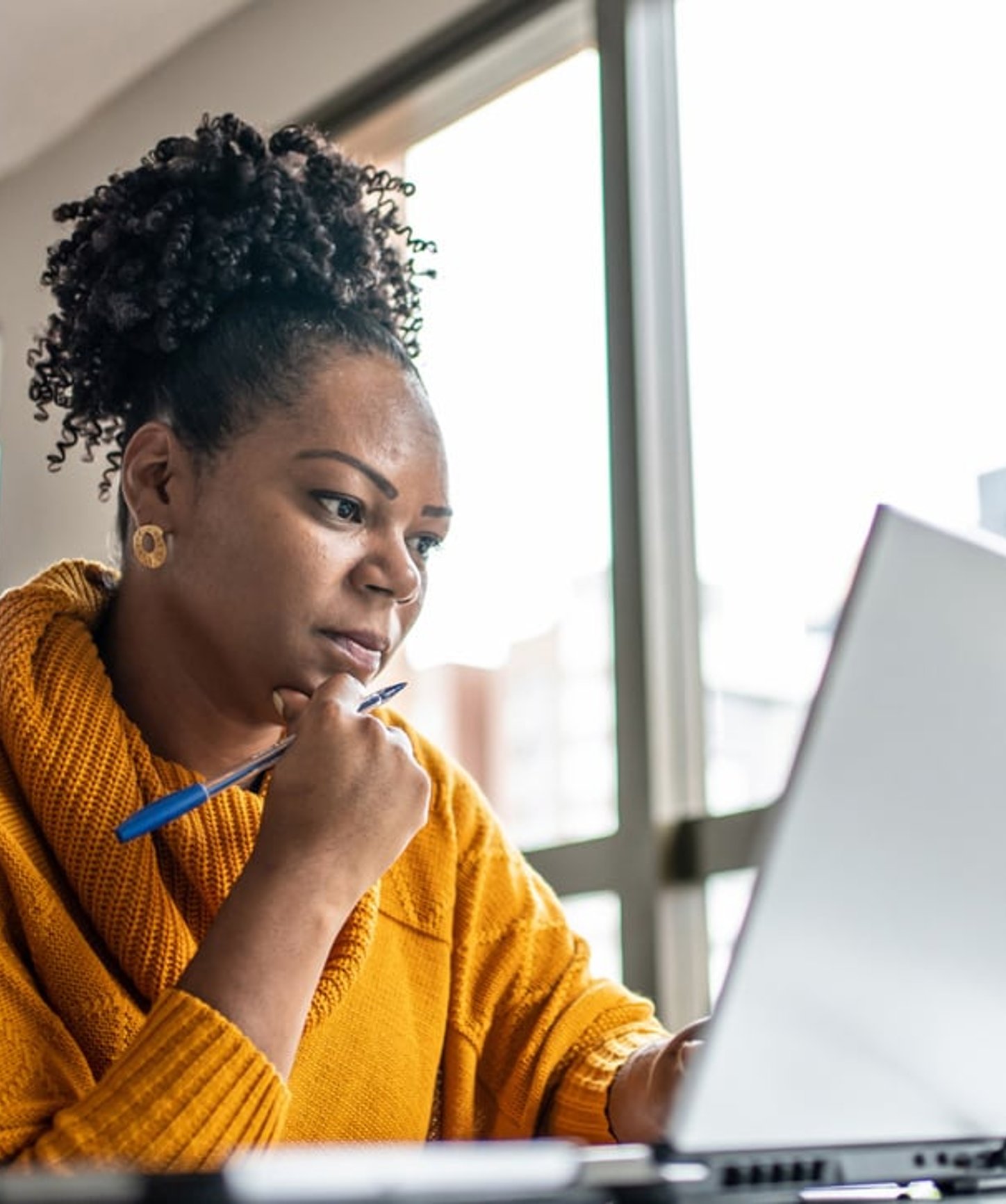 Focused woman in a mustard sweater working on a laptop, holding a pen while thinking in a bright modern workspace