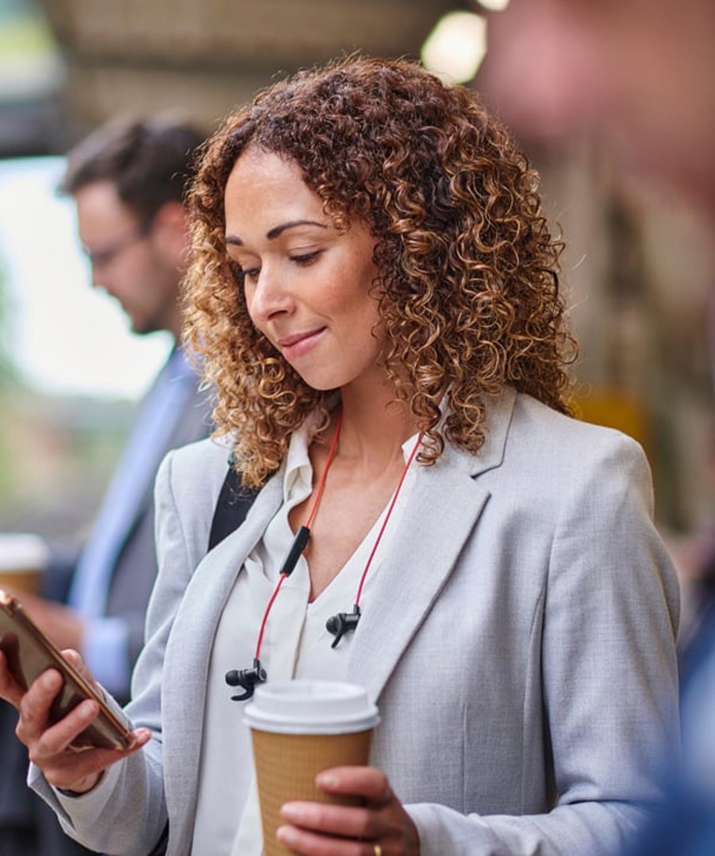 A woman in business attire stands at a train station, holding a coffee and looking at her phone with earphones around her neck, while commuters move in the background.