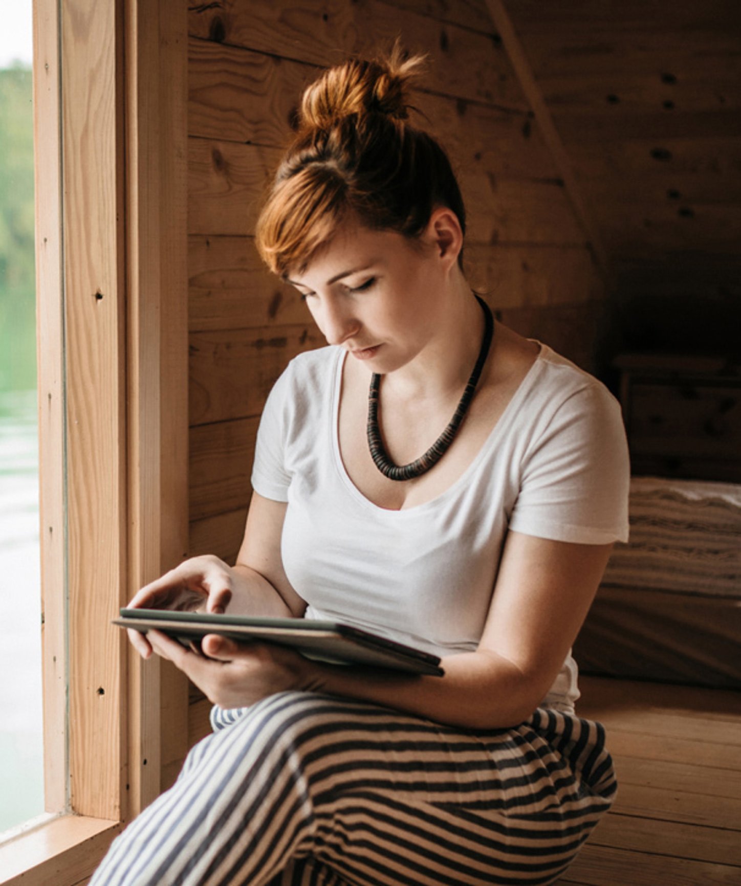 Young woman sitting by a wooden cabin window, reading on a tablet with a serene lakeside view in the background